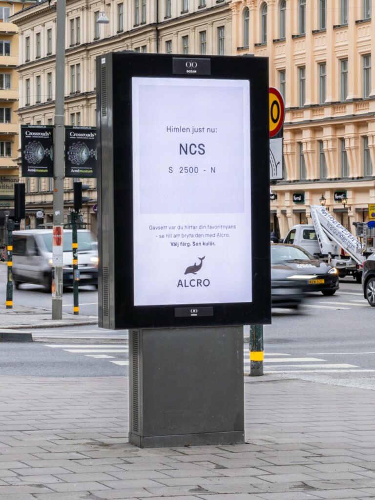 A digital out-of-home (DOOH) display on a sidewalk in an urban setting features an interactive AdTech campaign for Alcro. The screen displays the text “The Sky Right Now” along with a specific NCS color code and Alcro’s logo. 