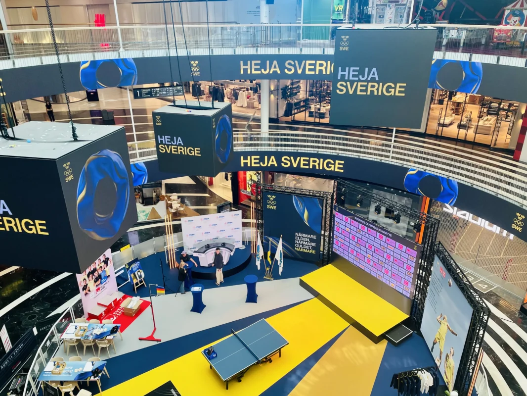 Översiktsbild av en eventyta och fanzone för Sveriges Olympiska Kommitté (SOK) med temat "Heja Sverige" i ett köpcentrum. Visar en uppbyggd scen med LED-skärm, stora hängande kuber, bordtennisbord och profilering i gult och blått.