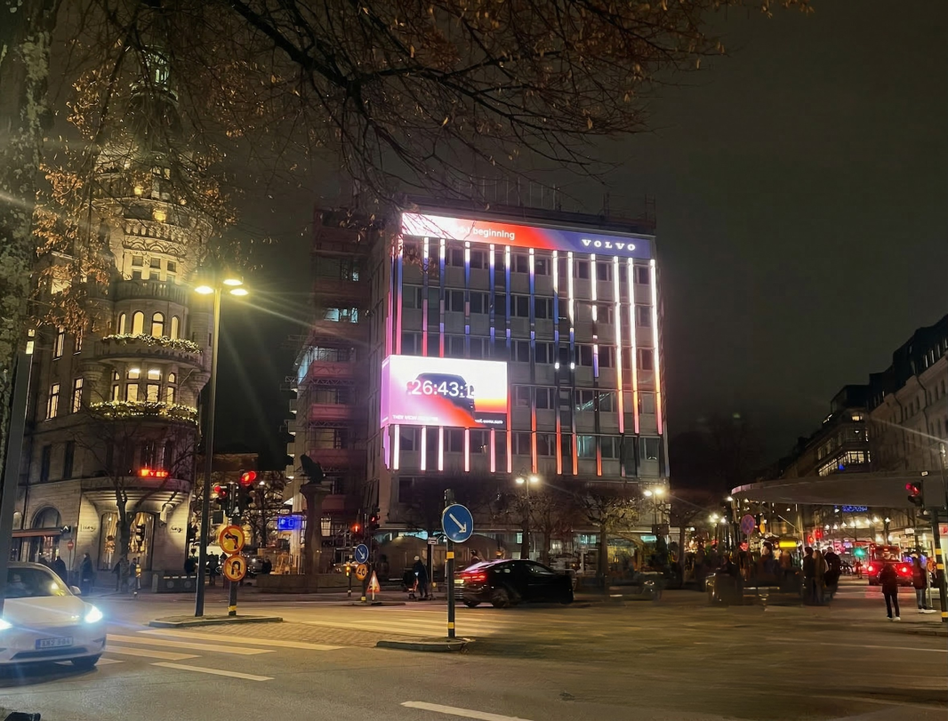 En stor digital billboard på en pampig byggnadsfasad i Stockholm i kvällsljus visar en nedräkning i realtid för Volvo EX60. Skärmen presenterar en röd timer bredvid bilens silhuett under Volvos logotyp, vilket illustrerar en storskalig och interaktiv utomhuskampanj.